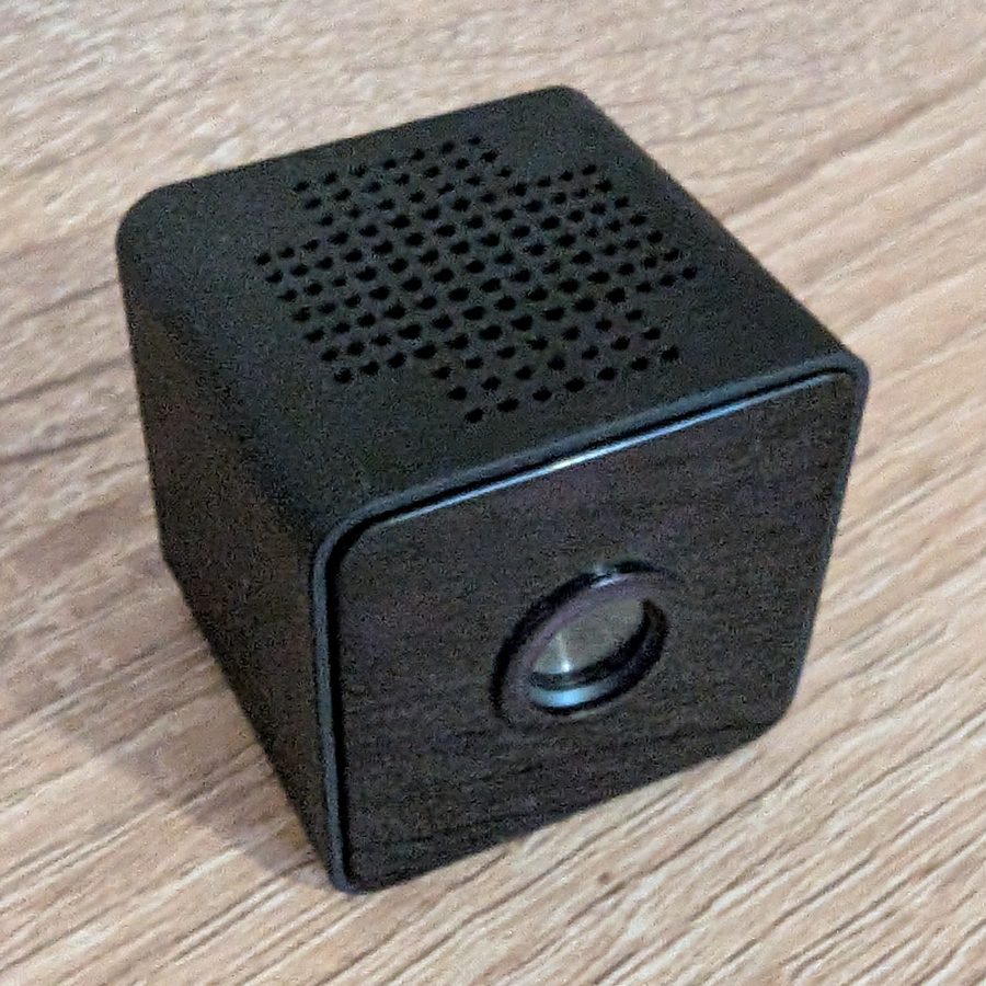 A black mini WiFi cube camera, about 3 cm wide, on a wooden desk